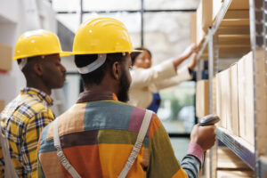 African american logistics managers scanning freight in storage department. Package handlers wearing safety hard hats searching customer order using barcode scanner in warehouse